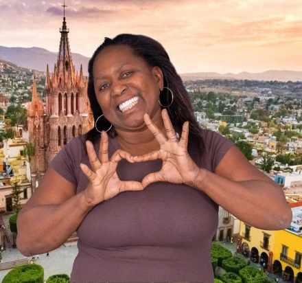 Smiling woman with brown skin forming a heart shape with her hands in front of her chest.