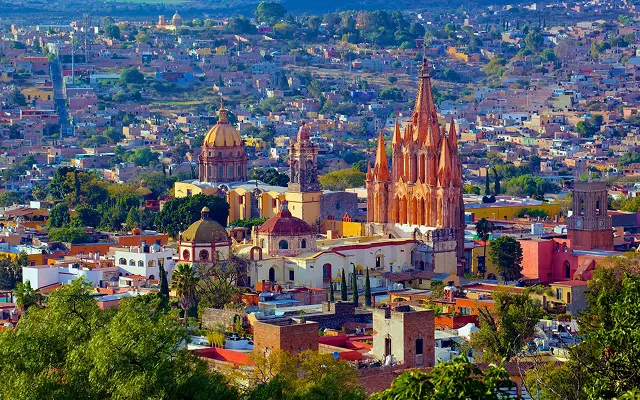 Distant view of San Miguel de Allende featuring its iconic baroque-style church surrounded by colorful colonial buildings and lush green trees.